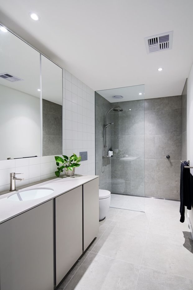 Bright contemporary bathroom with a long white vanity, integrated basin, large mirrored wall, toilet, and open walk-in shower. The room features white tiled walls, large light-grey floor tiles, grey stone-look shower wall tiles, recessed downlights, and clean minimal fixtures.
