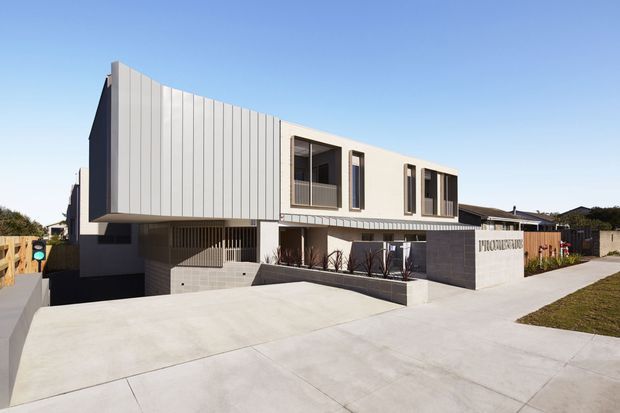 Contemporary multi-storey residential building viewed from the street, with a white vertically ribbed upper facade, dark lower masonry walls, large recessed windows, covered entry, and a broad concrete driveway. The clean geometric form and flat roof create a modern urban facade.