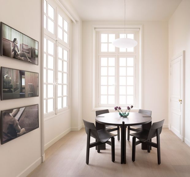 Bright, refined dining room with a round dark dining table and four dark sculptural chairs arranged beside tall multi-pane windows. White walls, white panelled doors and trim, pale timber flooring, a simple white pendant light, and framed artwork create an airy, understated interior.