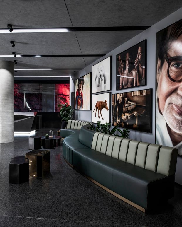 A dramatic, contemporary commercial reception or lounge interior with a long curved channel-tufted sofa in dark green and cream upholstery. The space features polished dark flooring, a textured concrete ceiling with linear strip lighting, a cylindrical concrete column, black geometric side tables, lush indoor plants, and a gallery wall of large framed photographic and artistic prints. A dark reception or bar-like area and red illuminated artwork are visible in the background.