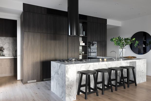 Contemporary open-plan kitchen featuring a large white-and-grey veined stone island with waterfall sides, dark timber-toned full-height cabinetry, integrated appliances, a black cylindrical rangehood, and four black bar stools. Light timber flooring, white walls, recessed ceiling lights, and a round black-framed wall mirror complete the refined, high-contrast interior.