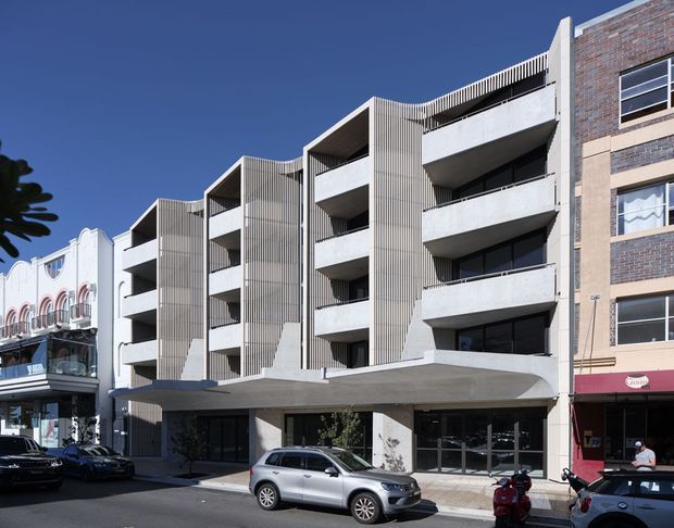 Street-level exterior view of a contemporary multi-storey apartment building in an urban commercial streetscape. The facade combines exposed concrete balcony slabs, vertical metal screening, recessed balconies, and a contrasting brick-clad side wall. Ground-floor glazing opens onto the street, with neighbouring retail and apartment buildings visible on either side.