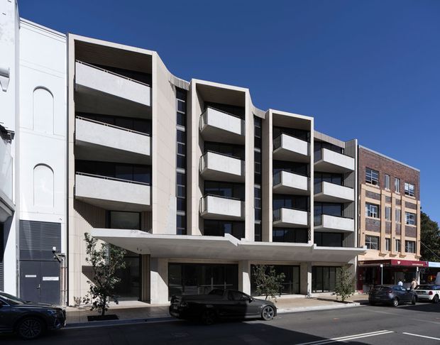 Street-level exterior view of a contemporary multi-storey apartment building in an urban setting. The facade combines pale rendered or concrete surfaces, projecting curved-front balconies, dark vertical window bands, and a recessed ground-floor entry or commercial frontage. An adjacent brick-and-masonry building is visible to the right.