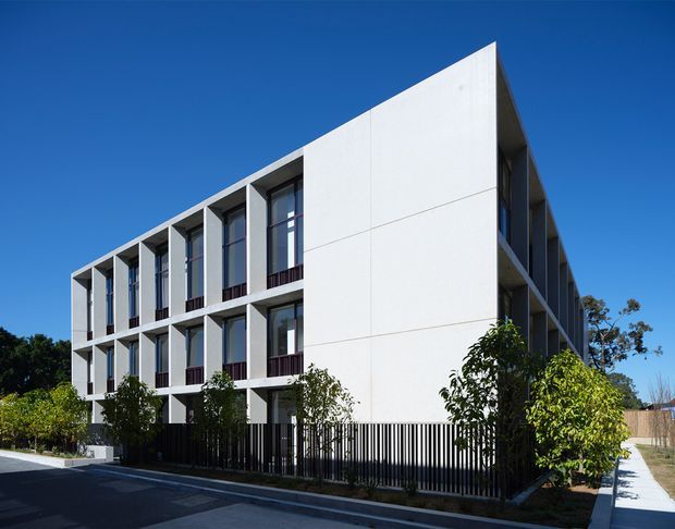 Exterior architectural photograph of a contemporary multi-storey residential building with a strong geometric form, white panelled facade, recessed floor-to-ceiling windows, narrow projecting balconies, and a black vertical metal fence. The building sits along a paved street and footpath under a clear blue sky, with landscaping around the frontage.