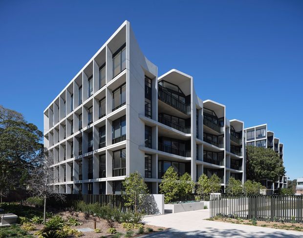 Exterior architectural photograph of a contemporary multi-storey apartment building. The building has a strongly articulated facade with repeated projecting balcony bays, deep recessed window openings, dark metal balcony balustrades, pale grey concrete or rendered walls, and a flat roofline. Landscaping, pedestrian paving, fencing, and mature trees surround the development under a clear blue sky.