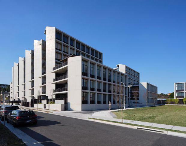 Exterior street view of a large contemporary multi-storey apartment complex with repetitive balconies, deep window reveals, pale concrete or rendered facades, and strong rectilinear massing. The development is set along a paved suburban street with landscaped lawn, footpaths, and minimal planting under a clear blue sky.