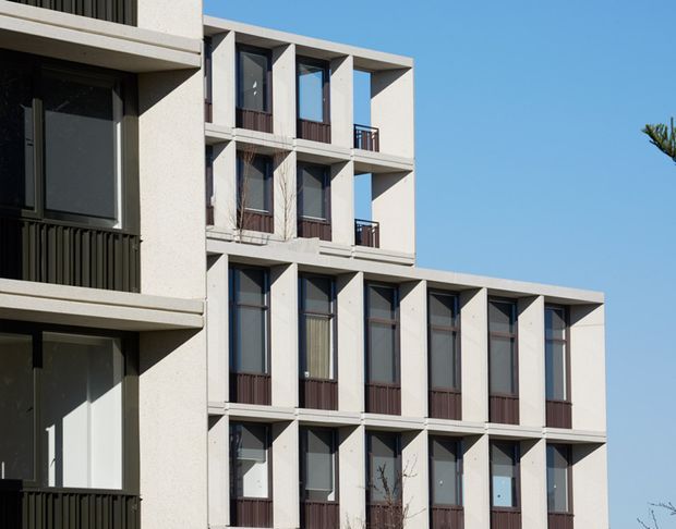 Exterior view of a contemporary multi-storey apartment building with a stepped massing profile. The pale rendered facade is structured by deep rectangular window bays, tall dark-framed glazing, projecting concrete slabs, and small dark metal balcony railings. Clear blue sky and minimal surrounding context emphasize the geometric facade composition.