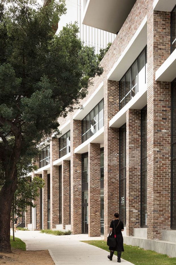 Exterior architectural photograph of a multi-storey brick building along a landscaped pedestrian walkway. The facade features repetitive tall brick piers, deep recessed entries, large dark-framed windows, projecting white horizontal balcony or canopy slabs, and a clean contemporary institutional or apartment-building composition. Mature trees line the walkway and a person provides scale.