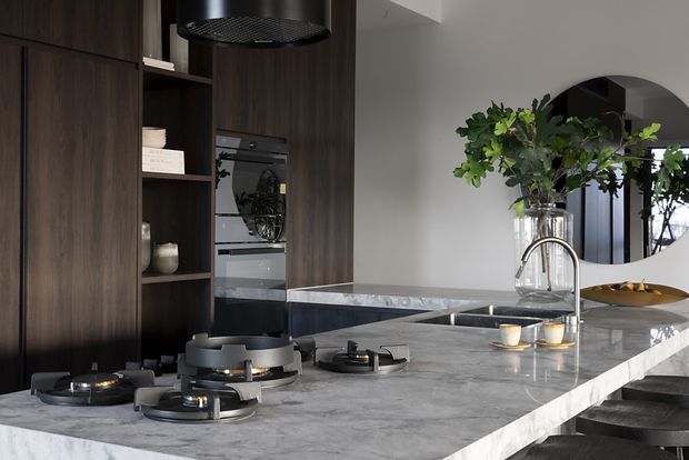 A contemporary kitchen dominated by a large grey stone island with an integrated gas cooktop and undermount sink. Dark timber cabinetry and open shelving conceal built-in ovens along the rear wall, while a black cylindrical rangehood hangs overhead. A curved chrome tap, bar stools, pendant-style rangehood, large arched mirror or opening, and restrained styling complete the sophisticated space.