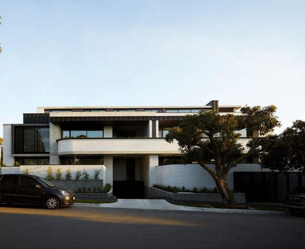 Street-facing photograph of a contemporary multi-storey residential building with a low, flat roofline, projecting balconies, dark metal screening, and pale small-format masonry cladding. The facade combines crisp horizontal bands with curved balcony edges and recessed entries, set behind a low wall and mature street tree.