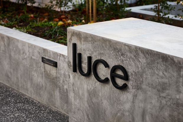 Close-up exterior view of a contemporary concrete rendered wall and raised entrance plinth, featuring black dimensional house lettering and a slim black wall light. Landscaping is visible behind the wall, with a dark textured driveway or path in the foreground.