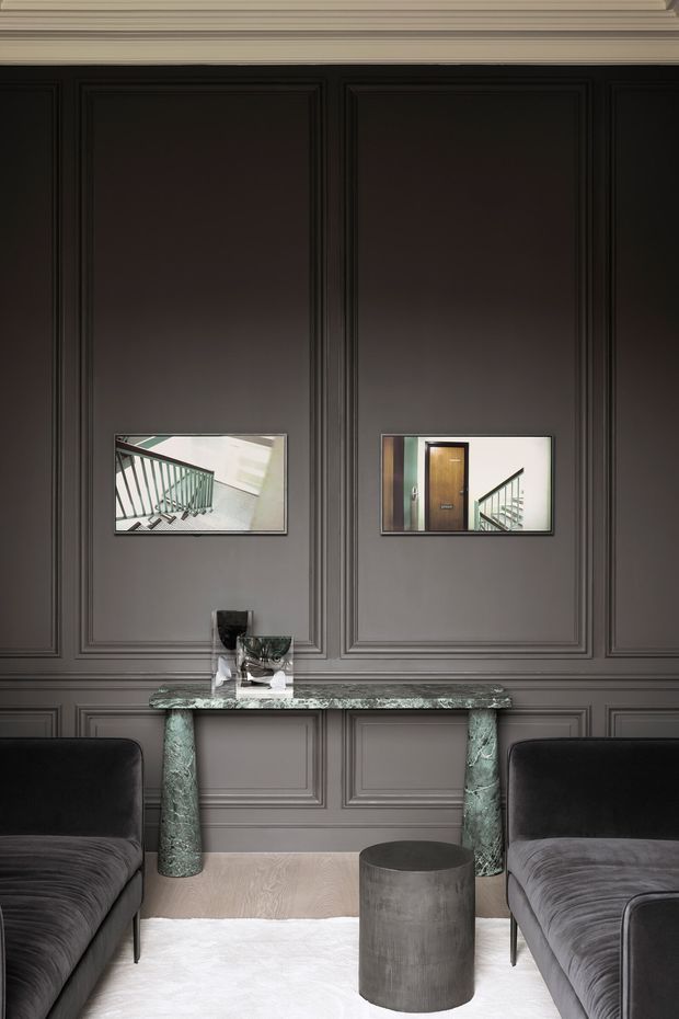 A refined, moody interior vignette featuring a narrow console table with green veined stone top and cylindrical stone legs, positioned against a dark grey wall with elaborate raised panel moulding and ornate ceiling trim. Two dark velvet sofas frame the foreground, with a small dark cylindrical side table and pale rug on the floor. Framed architectural images hang above the console.