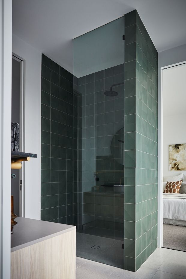A contemporary bathroom featuring a spacious glass-enclosed walk-in shower with dark green square wall tiles, matching tiled partition walls, dark shower fittings, and a grey tiled floor. A pale timber vanity is visible in the foreground, while an adjacent bedroom can be seen through the doorway.