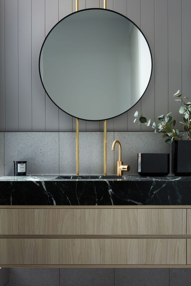 Close-up interior view of a refined bathroom vanity. A large round mirror with a thin black frame is mounted over vertically grooved dark grey wall panelling and a speckled grey stone backsplash. The vanity features a dramatic black veined stone benchtop, warm timber-grain cabinetry, and a sculptural brushed brass tap. Minimal black countertop accessories and a leafy plant add contrast.