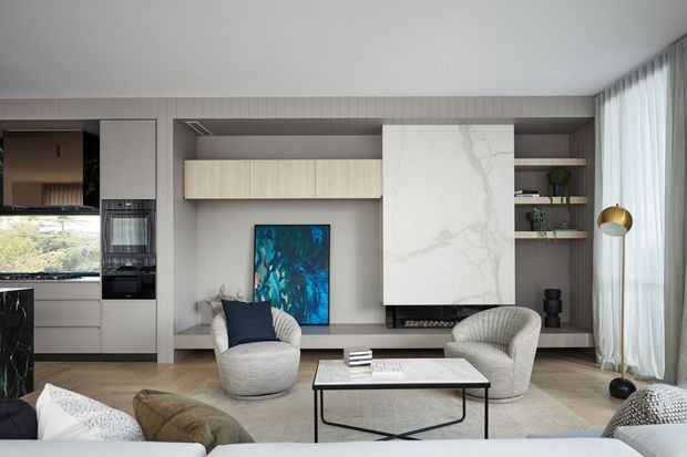 Contemporary living room with a refined neutral palette, featuring two sculptural upholstered lounge chairs, a low rectangular coffee table, a large abstract artwork, and a prominent veined stone fireplace surround. Built-in shelving and cabinetry frame the fireplace, while full-height sheer curtains bring in soft daylight. A compact kitchen with integrated appliances is visible at the left.