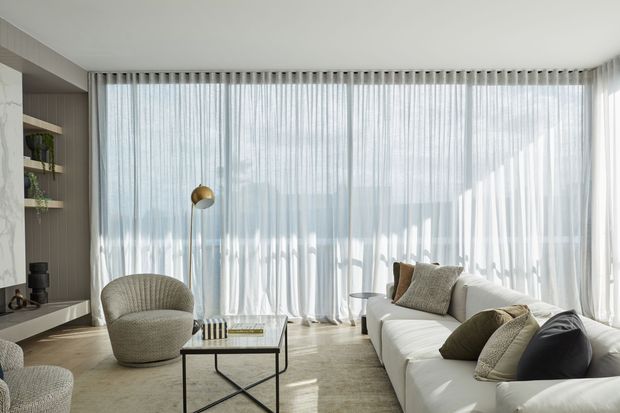Bright contemporary living room with a large white sectional sofa, textured armchair, black-framed glass coffee table, brass floor lamp, and full-height sheer curtains spanning a curved wall of glazing. A marble-clad fireplace or media wall and built-in shelving are visible at left, with a soft neutral palette and abundant diffused daylight.