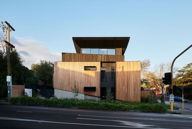 Street-facing photograph of a compact contemporary timber-clad house on an urban corner site. The building has a strongly rectilinear form, vertical wood cladding, a projecting upper-level volume with a broad flat roof, dark-framed openings, and a recessed central entry. Streetscape elements, landscaping, and utility infrastructure surround the facade.
