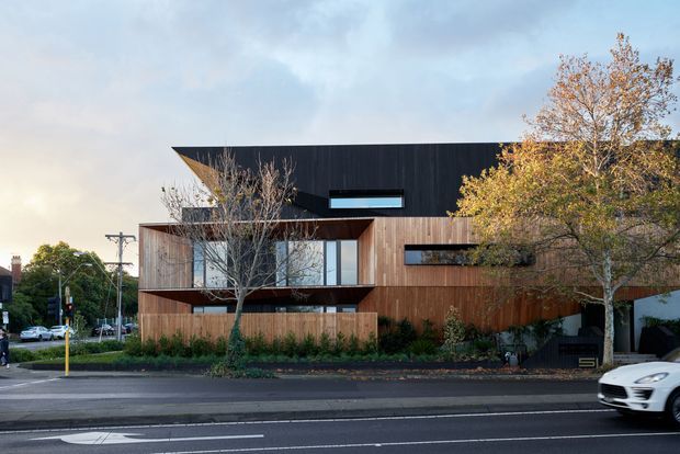 Street-facing view of a contemporary multi-level residence with a strongly horizontal composition, dark upper cladding, warm vertical timber-clad lower volumes, projecting cantilevered forms, and narrow horizontal windows. The building sits along an urban road with mature street trees and landscaped planting in front.