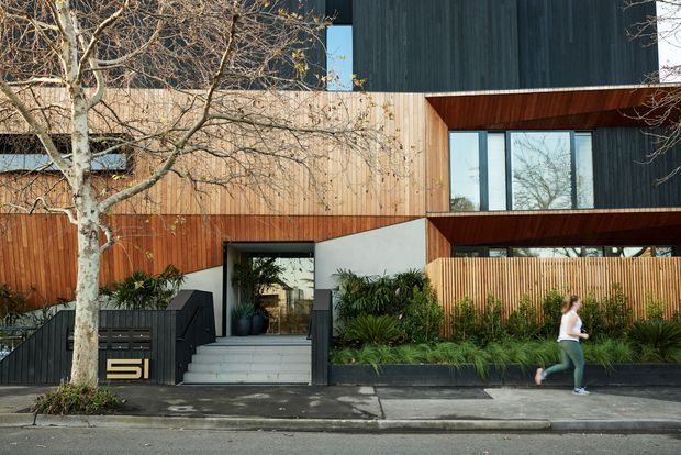Street-facing view of a contemporary multi-level residential building with dark vertical cladding, warm timber cladding and soffits, projecting box-like upper volumes, and large glazed openings. A recessed entry is reached by broad steps beside a dark slatted fence and landscaped planter beds. The image is photographed from the street and includes a pedestrian and mature street tree.