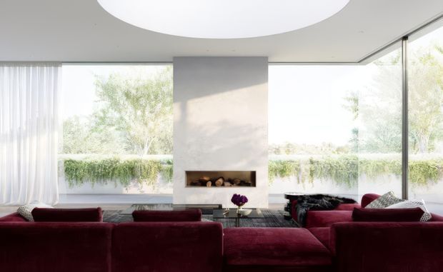 Bright, spacious contemporary living room with floor-to-ceiling glazing on two sides, sheer white curtains, a central pale stone or plaster fireplace wall with a long linear firebox, and deep burgundy upholstered sofas arranged around the fireplace. A large circular recessed ceiling feature or light adds a strong geometric element, while greenery is visible outside.