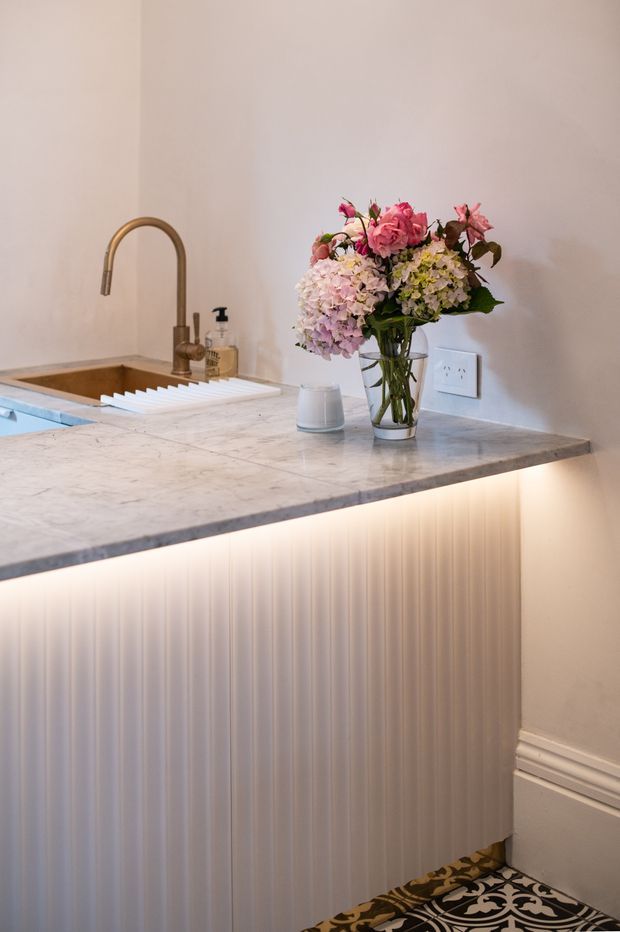 Close-up view of a refined kitchen island or peninsula with a pale veined stone benchtop, integrated warm under-bench lighting, vertical fluted panel cabinetry, and a brass-toned gooseneck tap beside a sink. A floral arrangement and soap dispenser sit on the bench, with decorative patterned floor tiles and traditional wall moulding visible at the edges.