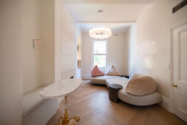 Bright, refined living room with a compact curved modular sofa arrangement, sculptural ottoman, pale textured upholstery, and a small organic-shaped console. The room features light herringbone timber flooring, warm off-white walls and built-in detailing, a decorative pendant light, and a tall window providing natural light.