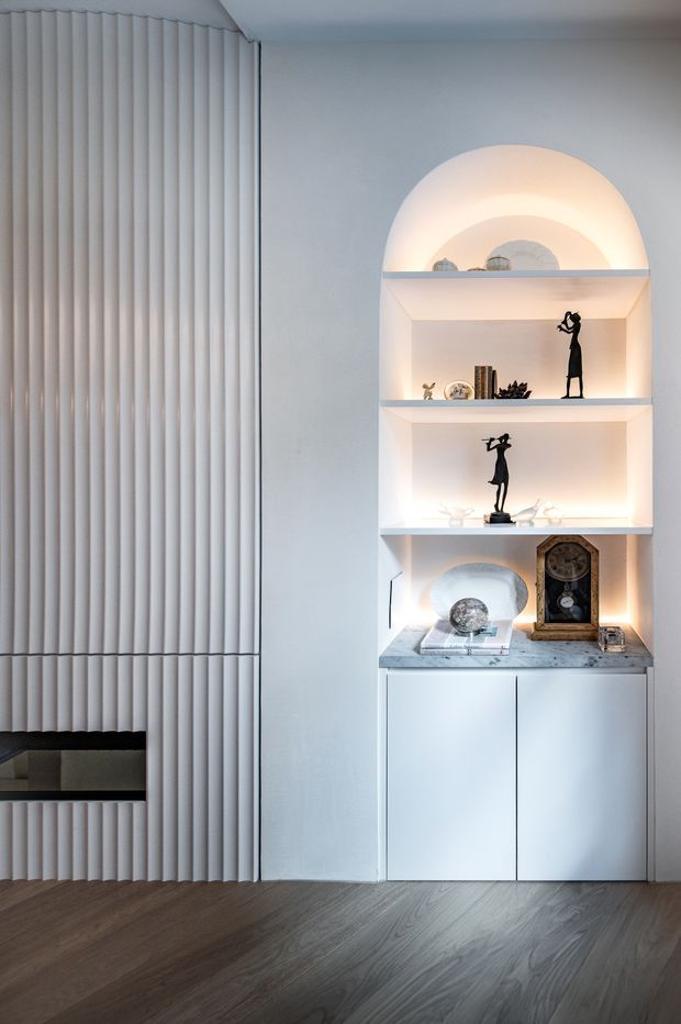 A refined interior feature wall with a full-height fluted vertical wall treatment and a built-in arched display niche. The illuminated open shelving contains decorative objects and framed collectibles above white lower cabinetry topped with a pale veined stone surface. Dark timber flooring contrasts with the predominantly white, softly lit joinery and wall surfaces.