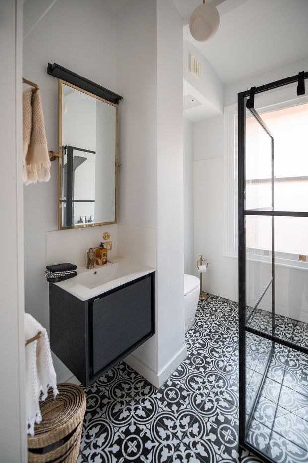 A bright, narrow bathroom with a black-and-white patterned tile floor, white walls, and a compact wall-mounted dark vanity with a white integrated basin. A brass-framed mirror hangs above the vanity, with a black wall-mounted light fixture and brass tapware. The toilet and a black-framed glass shower enclosure are visible at the rear, along with woven baskets and neutral towels. The room has a clean, graphic, contemporary character with strong monochrome contrast and warm metal accents.
