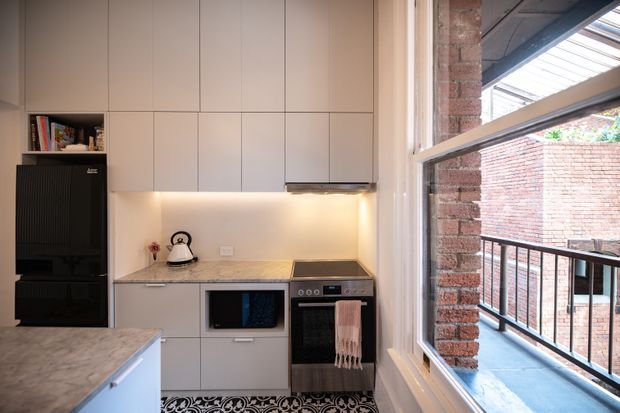 Compact contemporary kitchen with pale flat-panel cabinetry, a light stone benchtop, integrated appliances, black freestanding refrigerator, under-cabinet lighting, and a patterned dark floor mat. A large window reveals exposed brickwork and a balcony railing outside, bringing strong natural light into the space.