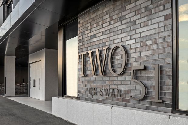 A modern commercial or hotel entrance corridor featuring a grey brick feature wall with large raised metallic lettering, dark metal soffit and window frames, pale stone or concrete plinths, and minimalist white service doors. The sheltered walkway has a refined, monochromatic contemporary appearance.