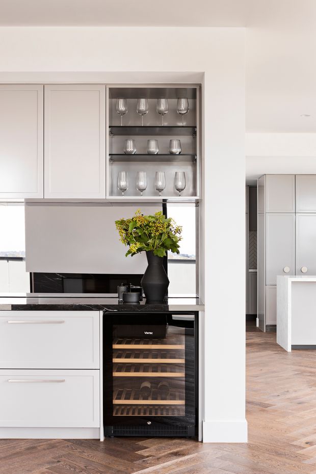 A bright, refined kitchen interior featuring pale grey-white framed cabinetry, a dark polished stone benchtop and splashback, illuminated glassware display shelving, and an integrated wine refrigerator. A black vase with flowers sits on the benchtop. Herringbone timber flooring continues into the adjacent space, where additional cabinetry and a white island are visible.