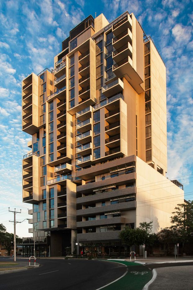 Exterior architectural photograph of a tall contemporary multi-storey apartment building in an urban setting. The building features projecting balconies, extensive dark-framed glazing, warm light-toned facade panels, vertical fins, and darker horizontal podium bands. A city street with marked bicycle infrastructure is visible in the foreground beneath a partly cloudy blue sky.