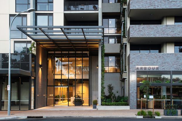 Street-level exterior view of a contemporary multi-storey apartment or mixed-use building. The facade combines large glazed panels, pale rendered wall sections, dark metal framing, projecting balconies, and light brick balcony cladding. A dramatic glazed entrance is sheltered by a broad steel-and-glass canopy, with landscaped planters and a ground-floor storefront visible at right.