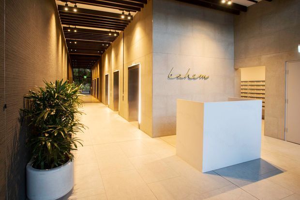 A refined commercial reception and corridor interior with a minimalist white reception desk, warm beige tiled flooring and wall panels, dark exposed ceiling beams with linear lighting, recessed doorways, and a large potted plant. A branded wall sign and lift or service doors suggest a hotel, office, or apartment lobby.