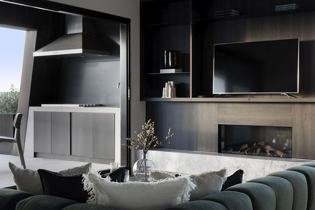 A sophisticated open-plan living room with a dark built-in media wall, flat-screen television, recessed fireplace, and open shelving. A concealed or adjoining kitchen is visible on the left with a stainless-steel rangehood, dark splashback, and minimalist cabinetry. The foreground features a large curved, channel-tufted sofa in a dark teal tone with layered neutral and black cushions. The room has a dramatic, refined palette of charcoal, black, grey, and off-white, with strong built-in joinery and a contemporary luxury feel.