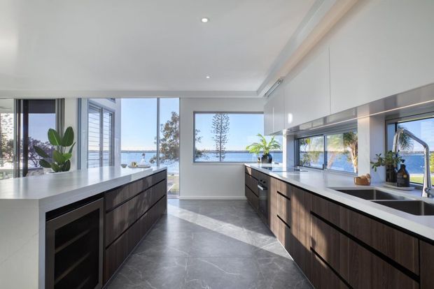 Bright contemporary kitchen with two parallel islands and extensive dark timber cabinetry, white stone benchtops, integrated appliances, and a large stainless-steel sink. Floor-to-ceiling glazing opens views toward a waterfront or coastal setting, while abundant natural light and restrained detailing create an airy, refined atmosphere.
