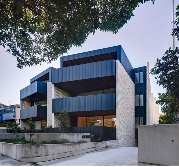 Exterior view of a contemporary multi-storey residential apartment building, featuring projecting balconies with dark blue-grey vertical metal cladding, pale brick masonry walls, recessed glazing, dark soffits, and exposed concrete retaining walls. The building is set along a landscaped sloping driveway and partly framed by mature trees.
