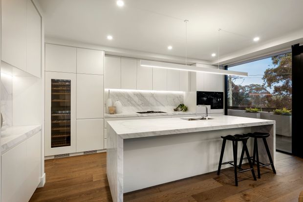 Bright contemporary kitchen with extensive white full-height cabinetry, a large marble-look island with integrated seating, a matching splashback, built-in wine fridge, concealed appliances, timber flooring, and a slim linear pendant light. Floor-to-ceiling glazing at the side provides views of trees outside.