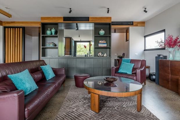 A contemporary living room with deep burgundy leather sofas and armchairs arranged around a round glass-topped timber coffee table. The room features dark built-in cabinetry with open shelving, integrated display storage, timber wall accents, a large horizontal window, polished concrete flooring, and turquoise cushions providing a vivid accent. Track lighting and restrained furnishings create a refined, modern interior.