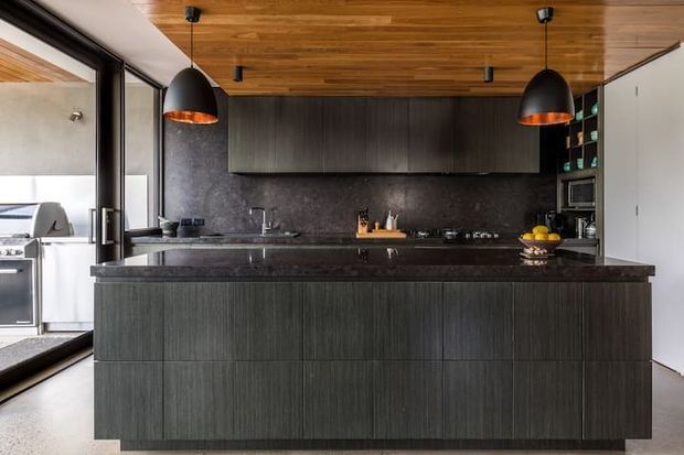 A sophisticated contemporary kitchen dominated by a large central island with dark vertically grained cabinetry and a dark stone benchtop. Matching dark cabinetry and a stone backsplash line the rear wall, while a warm timber ceiling adds contrast. Black pendant lights hang above the island, and full-height glazing or doors open toward an adjacent outdoor cooking area. Integrated appliances and open shelving are visible at the right.