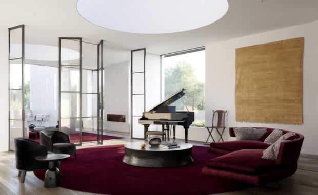 A refined contemporary living room with a grand piano, deep burgundy sectional sofa and rug, black lounge chairs, and a low round coffee table. Full-height black-framed glazed partitions create a bright, open connection to adjoining spaces, while a large circular ceiling light and textured wall artwork add strong focal elements.