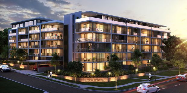 Architectural rendering of a contemporary multi-storey apartment or residential development at dusk. The complex has several connected blocks with flat roof overhangs, extensive floor-to-ceiling glazing, projecting balconies, warm interior lighting, dark vertical and horizontal facade elements, landscaped entrances, pedestrian paths, and an adjacent street with parked cars.