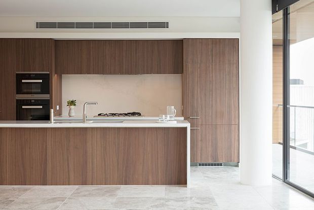 A contemporary minimalist kitchen with full-height dark timber-grain cabinetry, integrated black ovens, a pale stone island benchtop, gas cooktop, undermount sink, and concealed appliances. Large glazed doors open to a bright exterior, while pale stone-look flooring and white walls create a clean, refined atmosphere.