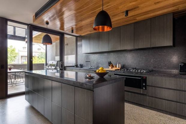 A contemporary kitchen with a large dark island, integrated cabinetry, stone benchtops, and a full-width cooking wall. The room features a warm timber-lined ceiling, black-framed glazing and doors opening to an outdoor dining area, dark pendant lights, and polished concrete or terrazzo-style flooring. The palette is predominantly charcoal, dark timber, black, and warm brown, with strong natural light entering from the left.