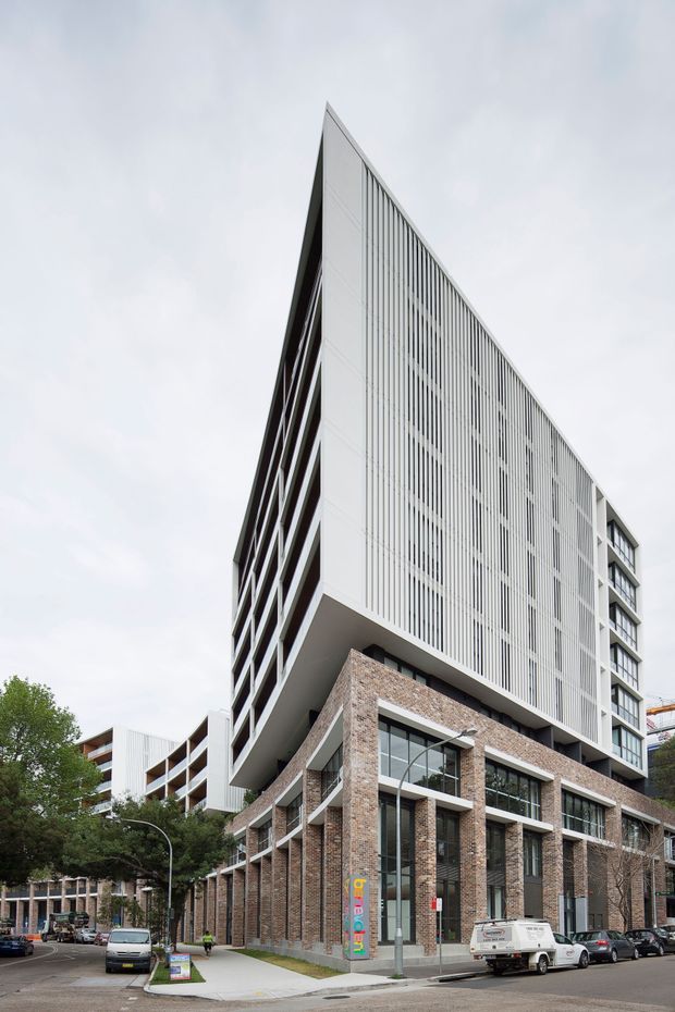Street-level exterior photograph of a contemporary multi-storey urban building. The lower levels feature a warm brick facade with tall glazed openings and projecting concrete framing, while the upper volume is sharply cantilevered and clad in pale vertical fins or louvres. Additional apartment or office wings are visible behind, with mature street trees and parked vehicles at ground level.