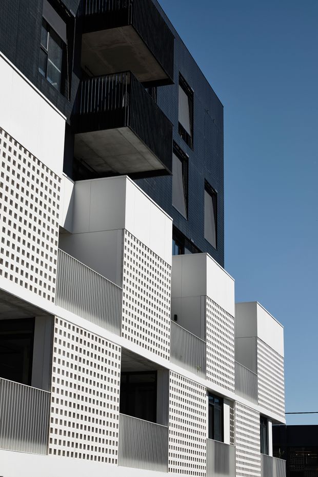 A close-up exterior architectural view of a contemporary multi-storey residential building. The facade combines dark blue-black tiled cladding with crisp white projecting volumes, perforated breeze-block screens, slim vertical metal balustrades, and deep concrete balconies. Repeated balconies and windows create a strong geometric rhythm against a clear blue sky.