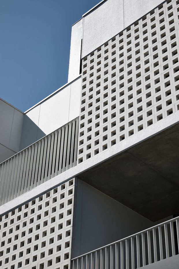 Upward-looking close architectural view of a modern multi-level building facade. The composition features white perforated concrete block screens, vertical metal balustrade or louvre elements, smooth light-coloured wall panels, and a projecting raw concrete slab against a clear blue sky. Strong geometric repetition, sharp shadows, and layered cantilevered forms create a clean, contemporary exterior.