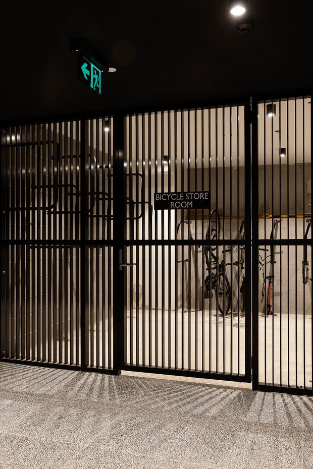 A dimly lit architectural interior entrance viewed through tall black metal security bars. A sign identifies a bicycle storage room, with several bicycles visible inside against pale walls and a light-coloured floor. An illuminated green emergency exit sign hangs above, while the bars cast strong linear shadows across the textured flooring, creating a utilitarian and high-contrast atmosphere.