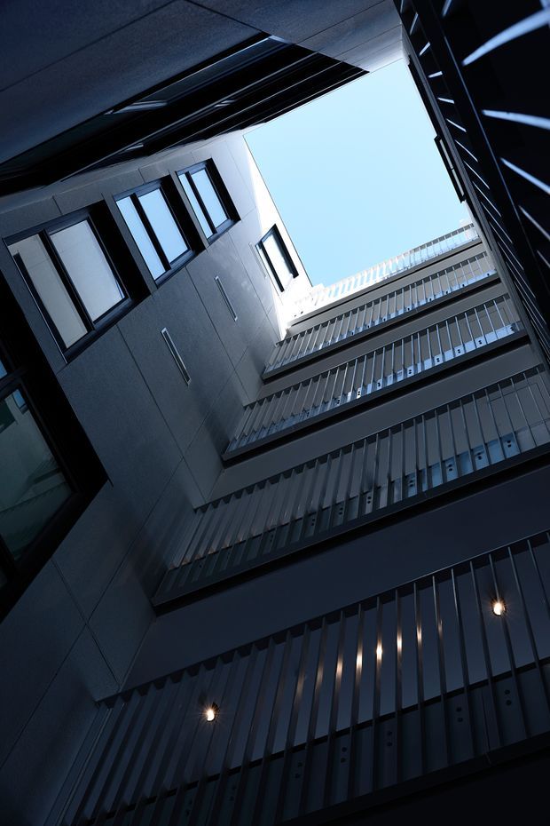 Upward-looking architectural photograph of a narrow multi-storey urban building atrium or courtyard. Dark metal-clad facades with repeated vertical fins, rows of windows, projecting balcony or circulation levels, and small wall lights frame a bright opening of sky. The image emphasizes strong vertical lines, geometric repetition, deep shadows, and a contemporary institutional or commercial character.