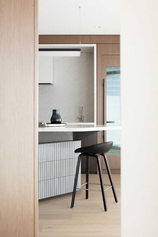 A refined contemporary kitchen viewed through a doorway, featuring warm timber cabinetry, a pale stone backsplash and benchtop, a compact island with vertically ribbed cabinetry, and a dark sculptural bar stool. A linear pendant light hangs above the island, with integrated appliances and a sink visible in the background. The space has a restrained, minimalist palette of timber, white, and black.